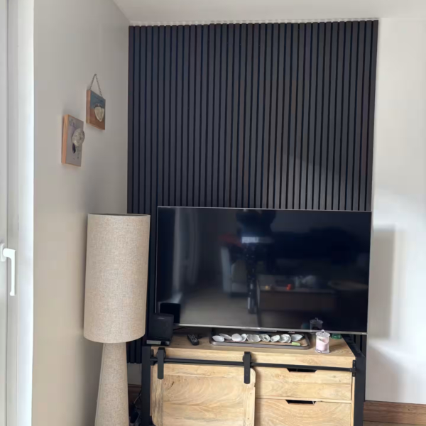Mur TV habillé du sol au plafond de panneaux acoustiques à lamelles chêne gris sur feutre noir, derrière un meuble TV en bois clair